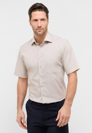 Uomo che indossa una camicia beige a maniche corte con bottoni e pantaloni scuri, in piedi davanti a uno sfondo bianco uniforme.