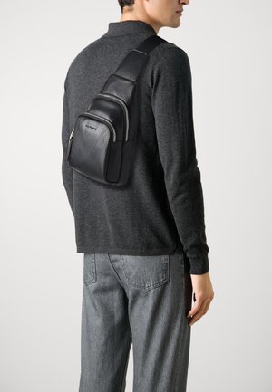 Sac à bandoulière en cuir noir avec une finition texturée et des zippers en argent, comportant trois compartiments, porté sur un pull en maille gris foncé.