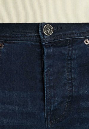 Tmavomodré denim džínsy majú uzatváranie na gombík a zips, s viditeľnými švíkovými detailmi a kovovými nitmi v blízkosti pásu.
