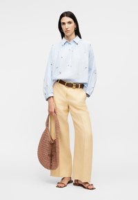 Femme portant un chemisier bleu clair brodé, un pantalon large beige, des sandales marron, tenant un sac à main tissé marron sur fond blanc.