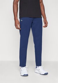 Mørkeblå joggebukser laget av bomullsmiksen, med en tettsittende design og elastisk midje, med en liten grønn logo på øvre lår.