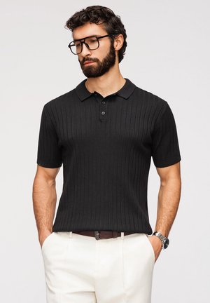 Mężczyzna o ciemnych kręconych włosach i brodzie, ubrany w czarną prążkowaną koszulkę polo, białe spodnie, czarny pasek, zegarek na rękę oraz czarne okulary na jednolitym tle.