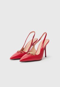 Zapatos de tacón alto de cuero rojo con tiras traseras y puntas afiladas, que presentan un detalle de cadena dorada en la parte frontal y delgados tacones de aguja.