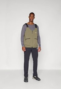 Man standing wearing olive green zip-up vest over long gray shirt, black pants, and black sneakers against plain gray background.