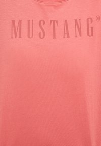 Tissu rose corail avec le mot "MUSTANG" imprimé dans une nuance légèrement plus foncée sur la zone de la poitrine.