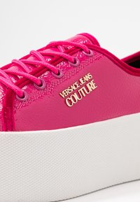 Sneaker rosa e lucida con una suola piatta bianca testurizzata, con il logo dorato "VERSACE JEANS COUTURE" e lacci rosa abbinati.