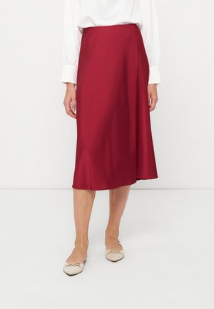 Femme portant une jupe rouge longueur genou, un chemisier blanc à manches longues et des chaussures plates beige avec un détail chaîne dorée.