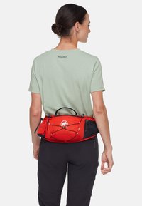 Borsa da vita rossa con tasca in rete e corde elastiche per lo stoccaggio, indossata sopra una maglietta verde chiaro, caratterizzata da un logo nero e un design funzionale.