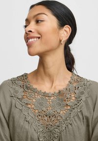 Blusa verde oliva con ricami intricati in pizzo floreale attorno al colletto. Sottile texture e vestibilità comoda. Orecchini a cerchio dorati visibili.