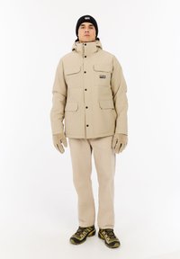 Beige vesivastainen takki, jossa on neljä etutaskua, kiinnitetty huppu ja mustat napit. Yhdistetty beigeihin housuihin ja ruskeisiin vaelluskengiin.