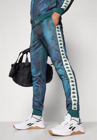 Chándal Kappa con un diseño de degradado azul-verde, franjas laterales blancas con detalles del logo, acompañado de una mochila negra y zapatos blancos de Reebok.