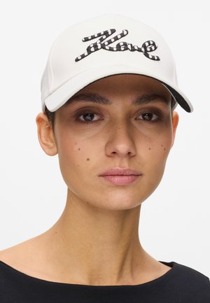 Γυναίκα που φοράει λευκό καπέλο του μπέιζμπολ με κεντητό κείμενο "Rave" σε μαύρες και λευκές ρίγες, συνδυασμένο με μαύρο τοπ και φυσικό μακιγιάζ.