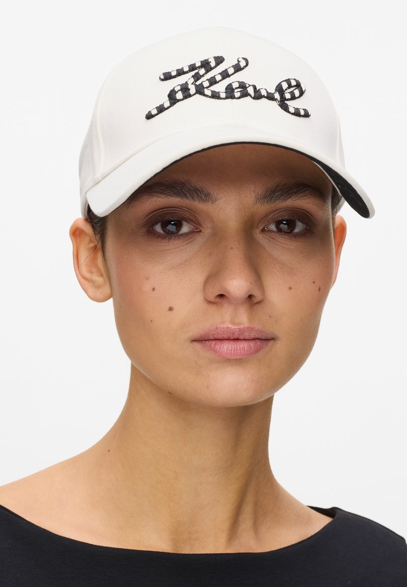 Γυναίκα που φοράει λευκό καπέλο του μπέιζμπολ με κεντητό κείμενο "Rave" σε μαύρες και λευκές ρίγες, συνδυασμένο με μαύρο τοπ και φυσικό μακιγιάζ.