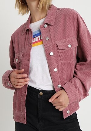 Persoon draagt een roze corduroy jasje over een wit T-shirt met een grafische opdruk en zwart, hooggetailleerd broek, die de voorkant van het jasje aanpast.