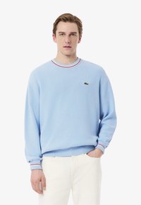Light blue pullover sweater with ribbed neckline featuring red and white stripes, soft texture, and a small Lacoste logo on the chest.