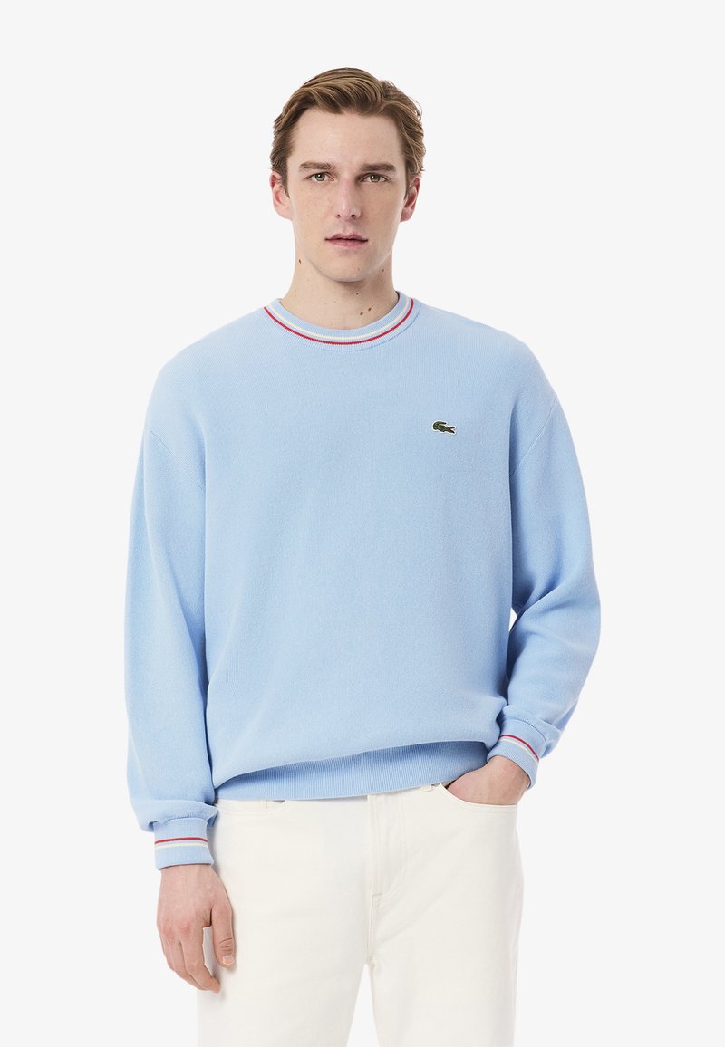 Maglione blu chiaro con scollatura a coste, caratterizzato da strisce rosse e bianche, texture morbida e un piccolo logo Lacoste sul petto.