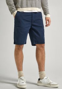 Shorts en coton bleu marine à coupe décontractée, dotés de poches latérales et d'ourlets cousus. Associés à des baskets blanches et des chaussettes côtelées crème.
