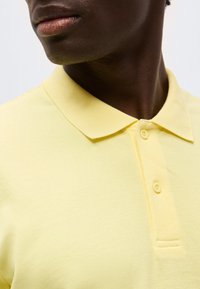 Κοντινό πλάνο ενός ατόμου που φοράει ανοιχτό κίτρινο πόλο πουκάμισο με γιακά και δύο κουμπιά κουμπωμένα.
