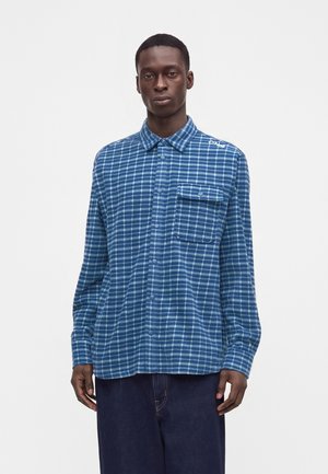Man met een blauw-wit geruit overhemd met borstzakflap, staand tegen een effen witte achtergrond.