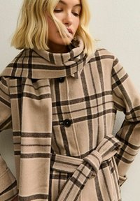 Kvinna iklädd en beige rutig kappa med matchande scarf, markerad midja och svarta knappar, står mot en enhetlig ljus vägg.