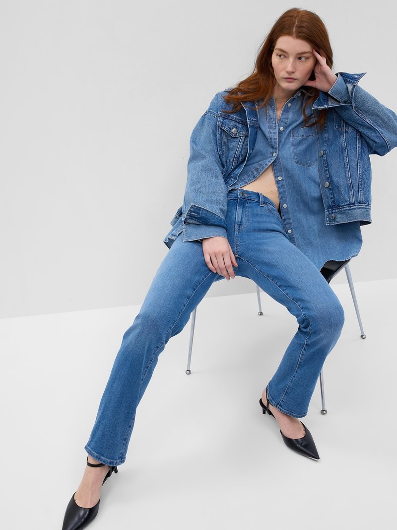 Chaqueta y camisa de mezclilla en azul claro, con mangas anchas y botones en el frente. Jeans ajustados azules y tacones negros de punta, sentada en una silla.
