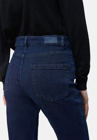 Jeans de tiro alto en azul oscuro con una textura suave, cinco bolsillos y un parche de cuero en la parte posterior. Diseño clásico de denim.