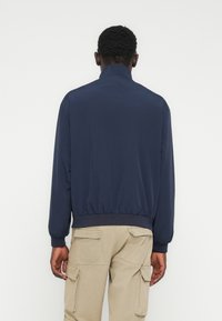 Άνδρας που φορ άει μπλε σκούρο μπουφάν και μπεζ παντελόνι cargo, στέκεται με την πλάτη στην κάμερα μπροστά σε ένα απλό λευκό φόντο.