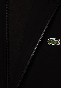 Giacca nera con zip, parzialmente aperta, che mostra un logo di coccodrillo verde ricamato sul tessuto vicino alla zip.