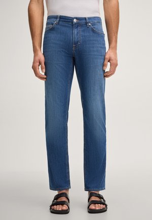 Mann trägt blaue Jeans mit geradem Bein, ein weißes Hemd und schwarze Sandalen mit zwei Riemen, steht vor einem schlichten Hintergrund.