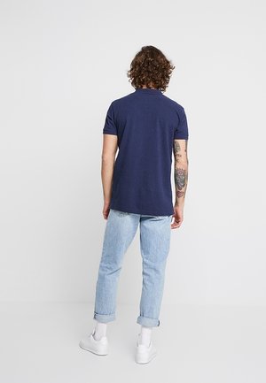 Navy short-sleeve polo shirt with a classic collar, paired with light blue rolled-up jeans and white sneakers. Features tattoos on the arm.