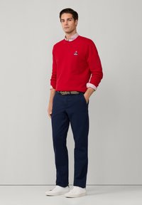 Sudadera de algodón roja con un pequeño logo, combinada con pantalones azul marino y zapatillas blancas. Un cinturón texturizado añade detalle al conjunto.