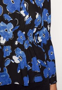 Floral blouse in black fabric with blue and white abstract flower patterns. Features gathered waist, long sleeves, and a loose fit.