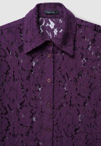 Maglietta di pizzo viola con motivo floreale, dotata di colletto e cinque bottoni marroni sul davanti. Tessuto leggero e trasparente.