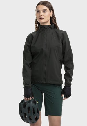 Schwarze, leichte Jacke mit hohem Kragen und Frontreißverschluss, kombiniert mit grünen Shorts und schwarzen Handschuhen. Die Person hält einen dunklen Helm.