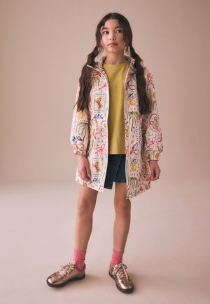 Pige med langt hår i fletninger iført en mønstret frakke, gul skjorte, denim nederdel, lyserøde sokker og metalliske sneakers, der står på en beige baggrund.