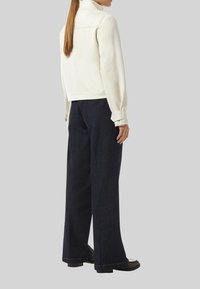 Biała zamszowa kurtka z kołnierzem, w połączeniu z ciemnoniebieskimi szerokimi spodniami i czarnymi butami, widziana od tyłu.