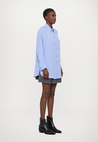 Camicia button-up blu chiaro con colletto, maniche lunghe e vestibilità rilassata, abbinata a una gonna grigia plissettata e stivali neri alla caviglia con dettagli.
