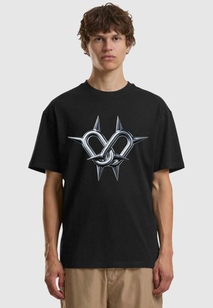 Schwarzes Baumwoll-T-Shirt mit einem metallisch silbernen, herzförmigen Design, das in der Mitte mit Schlaufen und Spitzen verwoben ist. Kurze Ärmel.