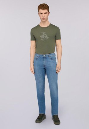 Camiseta ajustada de color verde oliva con un estampado gráfico, combinada con jeans de pierna recta de color azul claro y zapatillas de color verde oscuro, sobre un fondo liso.
