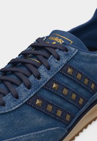 Κοντινό πλάνο μπλε αθλητικού παπουτσιού Adidas με μπλε σκούρα κορδόνια, χρυσά καρφιά στις πλαϊνές ρίγες και χρυσό λογότυπο Adidas στη γλώσσα.