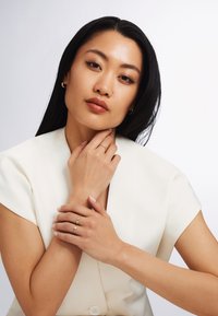 Model wearing a cream blazer displays gold rings on her fingers, featuring smooth and twisted designs, with a small pearl accent on one ring.
