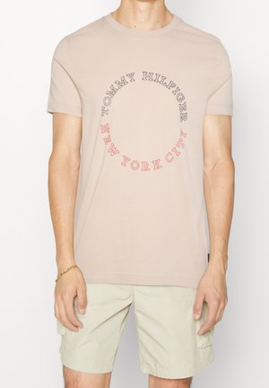 Mand iført beige Tommy Hilfiger T-shirt med cirkulær tekst "Tommy Hilfiger New York City" og lysebeige shorts.