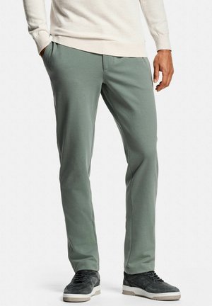 Man draagt lichtgroene rechte broek, zwarte sneakers en een lichtbeige trui met lange mouwen, staand met één hand in de zak.