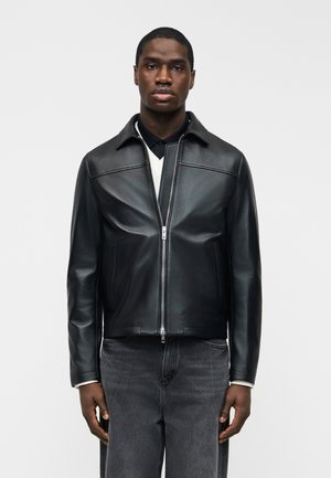 Άνδρας που φοράει μαύρο δερμάτινο μπουφάν με φερμουάρ πάνω από λευκό πουλόβερ και μαύρο πουκάμισο με γιακά, σε συνδυασμό με σκούρο γκρι τζιν, στέκεται μπροστά σε λευκό φόντο.