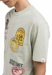 T-shirt in cotone grigio chiaro con toppe stampate colorate, tra cui motivi floreali e hawaiani, collo rotondo e maniche corte.