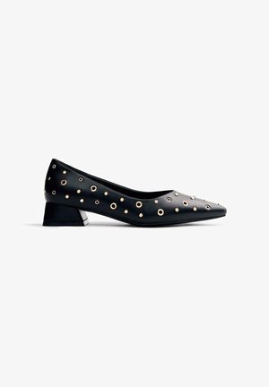 Escarpin en cuir verni noir avec un bout pointu et un petit talon carré. Accentué de grommets en métal doré de tailles variées sur le dessus.