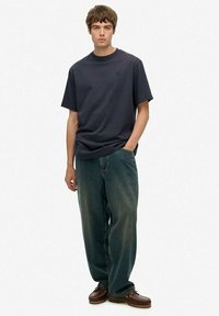 Jonge man die staat met één hand in zijn zak, gekleed in een donkerblauwe T-shirt, ruime vervaagde jeans en bruine leren schoenen.