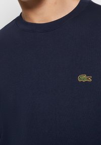Homme portant un t-shirt bleu foncé à col rond avec un petit logo crocodile vert brodé sur la poitrine gauche.