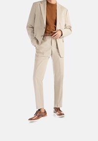 Completo beige chiaro con giacca aderente e pantaloni con pence, abbinato a una maglietta marrone e scarpe eleganti marroni con suole bianche.