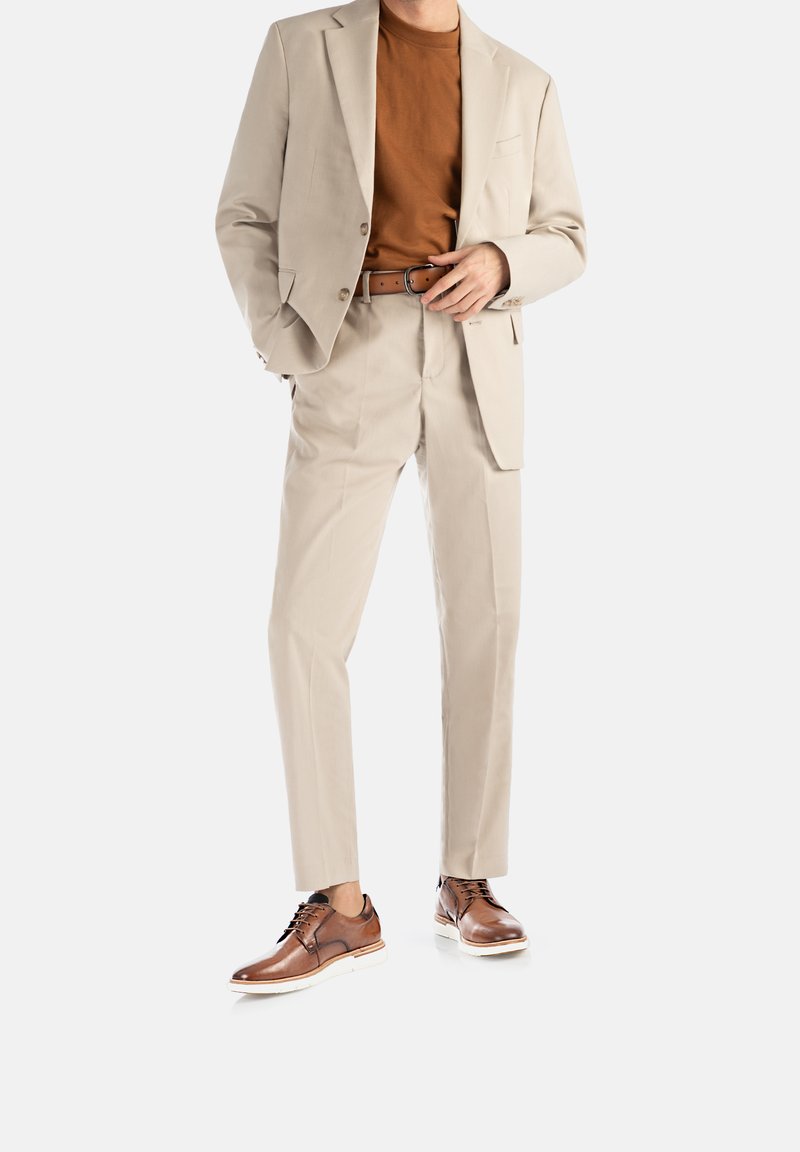 Completo beige chiaro con giacca aderente e pantaloni con pence, abbinato a una maglietta marrone e scarpe eleganti marroni con suole bianche.